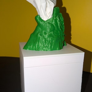 Pode incluir: Um porta-caixas de lenços de papel verde, texturizado, impresso em 3D, com um design orgânico estilizado. Os lenços brancos são puxados de cima. O suporte está sobre uma caixa retangular branca contra um fundo amarelo.