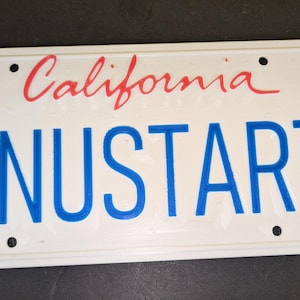 Puede incluir: Placa de matrícula blanca con la palabra "California" en escritura roja sobre las letras azules "ANUSTART". La placa de matrícula tiene cuatro orificios de montaje y un borde blanco.