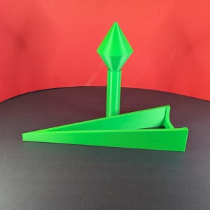 Puede incluir: Objeto verde brillante impreso en 3D con un diseño geométrico. Presenta un elemento vertical alto rematado con una forma de diamante y una pieza horizontal en ángulo. El objeto está sobre un fondo rojo.