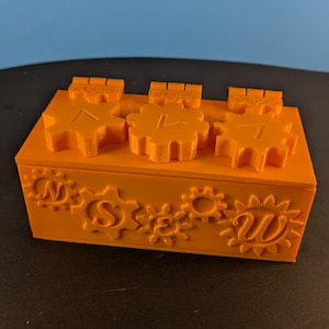 May include: An orange, rectangular box featuring gear-shaped designs and the letters N, S, and W. The top has three gear-shaped pieces with the letters A, L, and 7. The box is on a black surface.