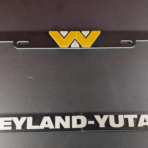 May include: Black license plate frame with a silver border and the text "WEYLAND-YUTANI" in white. The top features a yellow and white logo. The frame has mounting holes for easy installation.