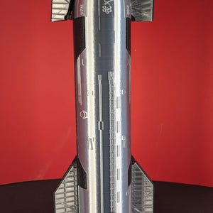 Peut inclure: Une maquette de fusée argentée et noire, avec des lignes de panneaux détaillées et des structures en forme d'ailes. La fusée a un cône de nez pointu et un corps cylindrique. Le modèle est posé sur un fond rouge.