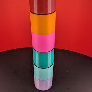 May include: A stack of six cylindrical objects in various colors: burgundy, orange, pink, teal, light blue, and lavender. The objects are stacked vertically against a red background.