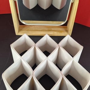 Può includere: Un organizer geometrico bianco con nove scomparti, riflesso in un piccolo specchio quadrato. L'organizer è realizzato in un materiale di colore chiaro e ha un design a forma di diamante. Lo specchio è incorniciato in legno chiaro.