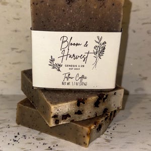 Puede incluir: Tres barras de jabón Tallow Coffee apiladas, con una etiqueta blanca alrededor de la barra superior. La etiqueta dice "Bloom & Harvest" con detalles florales y "Tallow Coffee". El jabón es de color marrón claro con motas oscuras.