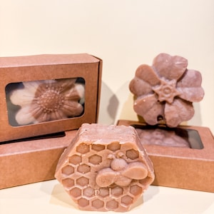 Puede incluir: Jabones artesanales en tonos marrón claro, con diversos diseños. La imagen presenta un jabón con patrón de panal y una abeja, un jabón con forma de flor y un jabón con forma de flor dentro de una caja marrón con ventana transparente. El fondo es amarillo claro.