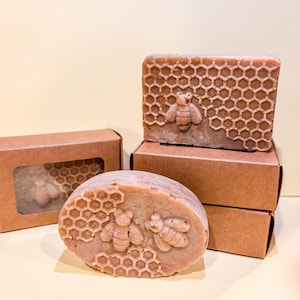 May include: Handmade soap bars with bee and honeycomb designs. One oval bar and one rectangular bar are visible, along with a rectangular bar in a box. The soap is light brown with detailed bee and honeycomb patterns.