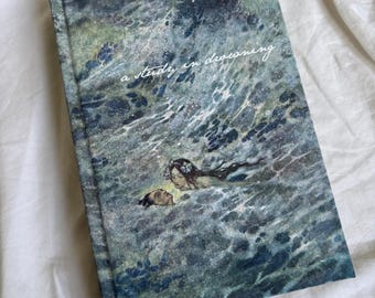 Puede incluir: Un libro de tapa dura con una ilustración en acuarela de figuras en el agua. La portada presenta el título "a study in drowning" en una escritura delicada. La obra de arte es predominantemente azul y verde, evocando una sensación de profundidad y misterio. El libro descansa sobre una superficie blanca y texturizada.