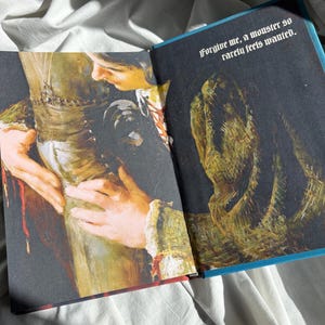 May include: An open book featuring an illustration of a person embracing a textured, dark green creature. The text printed in white at the top right corner reads, "Forgive me, a monster so rarely feels wanted." The artwork uses deep shadows and highlights.