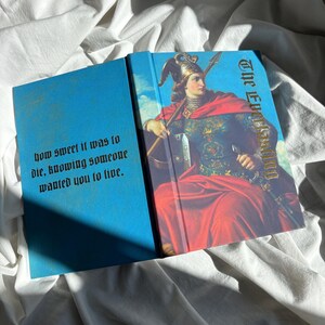 May include: A hardcover book with a bright blue cover featuring a painting of a figure in a winged helmet and red cloak holding a spear. The text on the front reads The Everlasting and the back cover text reads how sweet it was to die, knowing someone wanted you to live.