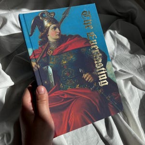 May include: A hardcover book titled The Everlasting featuring a classical painting of a figure in ornate armor and a red cloak holding a spear against a blue sky. The title is printed in a metallic gold gothic font on the front cover.