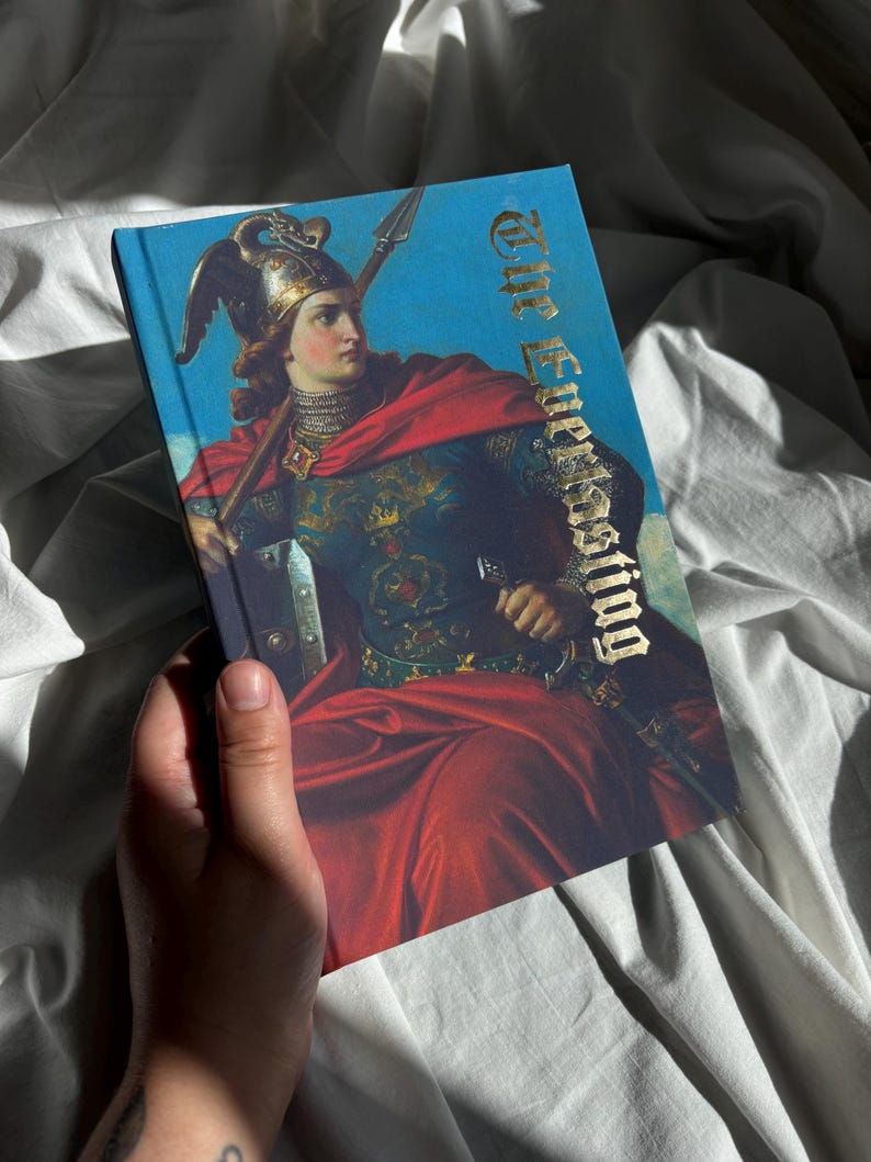 May include: A hardcover book titled The Everlasting featuring a classical painting of a figure in ornate armor and a red cloak holding a spear against a blue sky. The title is printed in a metallic gold gothic font on the front cover.