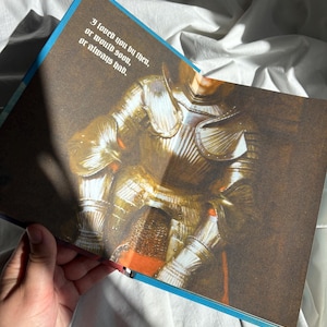 May include: An open book page featuring a detailed illustration of a suit of polished gold armor. The text printed on the page reads: I loved you by then, or would soon, or always had. The illustration shows metallic textures and highlights.