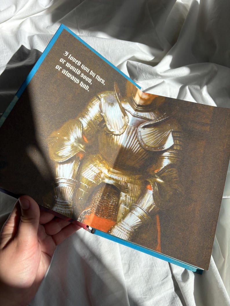 May include: An open book page featuring a detailed illustration of a suit of polished gold armor. The text printed on the page reads: I loved you by then, or would soon, or always had. The illustration shows metallic textures and highlights.