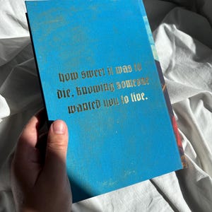 May include: A rectangular book with a bright blue cover featuring gold-colored text in a gothic font that reads: how sweet it was to die, knowing someone wanted you to live. The book has a smooth, matte finish and is held against a white fabric background.