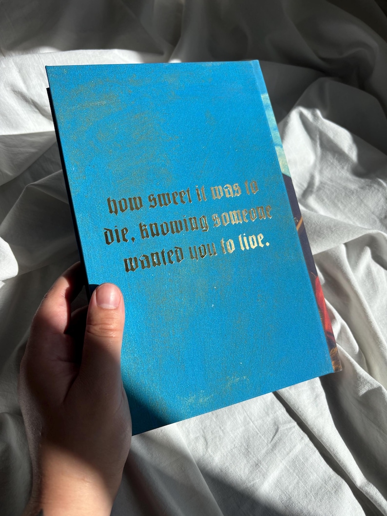 May include: A rectangular book with a bright blue cover featuring gold-colored text in a gothic font that reads: how sweet it was to die, knowing someone wanted you to live. The book has a smooth, matte finish and is held against a white fabric background.