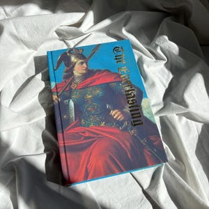 May include: A hardcover book titled The Everlasting featuring a vibrant illustration of a figure in medieval armor with a winged helmet and a red cloak, holding a spear and sword against a bright blue sky background with detailed gold lettering.