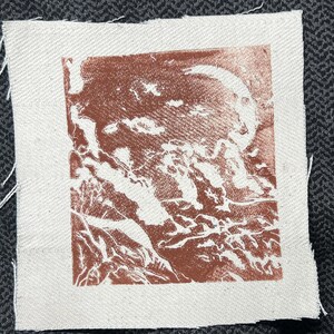 May include: A square, off-white fabric patch with a brown print. The print depicts a detailed, abstract landscape with a celestial body. The edges of the fabric are unfinished, with loose threads visible.