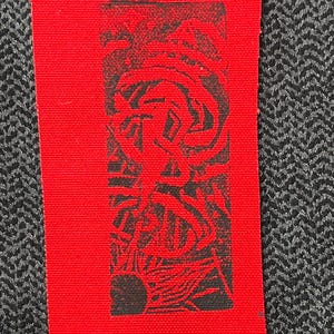 Puede incluir: Un parche de tela rojo rectangular con un diseño gráfico negro. El diseño parece ser una ilustración o símbolo abstracto. El parche se encuentra sobre un fondo gris oscuro texturizado.