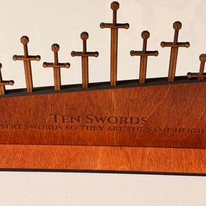 Może przedstawiać: Drewniana układanka z dziesięcioma mieczami o różnych rozmiarach. Miecze są wkładane w drewnianą podstawę z napisem "TEN SWORDS INSERT SWORDS SO THEY ARE THE SAME HEIGHT". Podstawa i miecze są w ciepłym brązowym kolorze.