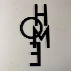 Op de afbeelding: Zwarte metalen wandkunst met het woord "HOME". De letters zijn in een modern, minimalistisch lettertype, waarbij de "O" een cirkel vormt. Het stuk is ontworpen om aan de muur te worden bevestigd en voegt een decoratief tintje toe aan elke woonruimte.