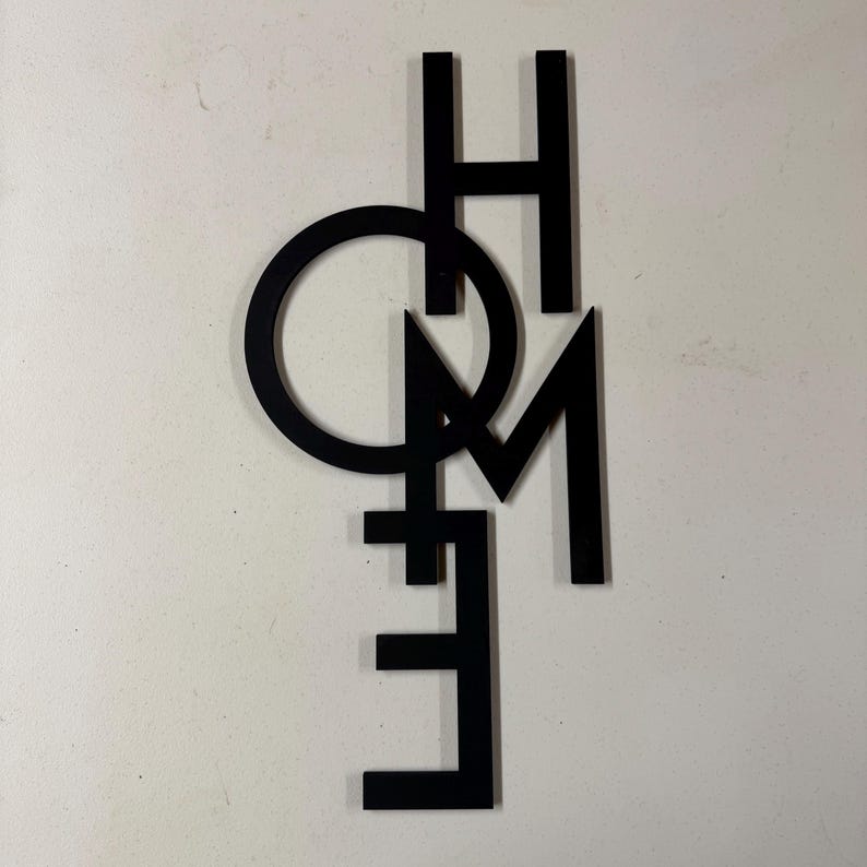 Op de afbeelding: Zwarte metalen wandkunst met het woord "HOME". De letters zijn in een modern, minimalistisch lettertype, waarbij de "O" een cirkel vormt. Het stuk is ontworpen om aan de muur te worden bevestigd en voegt een decoratief tintje toe aan elke woonruimte.