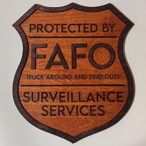 May include: Wooden shield-shaped sign with the text "PROTECTED BY FAFO (FUCK AROUND AND FIND OUT) SURVEILLANCE SERVICES" in dark brown lettering. The sign has a dark brown border and a light brown interior.