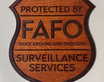 Può includere: Cartello in legno a forma di scudo con la scritta "PROTECTED BY FAFO (FUCK AROUND AND FIND OUT) SURVEILLANCE SERVICES" in lettere marrone scuro. Il cartello ha un bordo marrone scuro e un interno marrone chiaro.