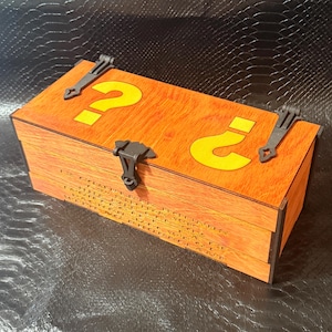 Può includere: Una scatola rettangolare in legno con finitura a venature color arancio chiaro, caratterizzata da due grandi punti interrogativi gialli sul coperchio. La scatola è fissata con cerniere e una chiusura centrale in metallo grigio scuro, con testo inciso.