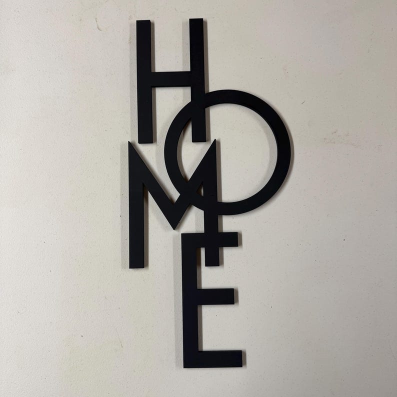 Op de afbeelding: Zwarte metalen wandkunst met het woord "HOME". De letters zijn in een modern, minimalistisch lettertype, waarbij de "O" de "M" en "E" overlapt. De kunst is gemonteerd op een lichtgekleurde muur.