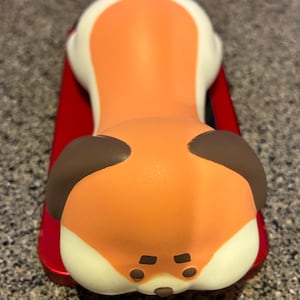 May include: A whimsical orange and white fox-shaped phone stand. The fox has brown ears and a cream-colored belly. The stand is resting on a red surface, creating a playful and functional accessory for any desk or workspace.