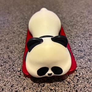 May include: A white and black panda figurine lying on a red rectangular base. The panda has black ears, eyes, and nose, with a white body. The figurine is a cute, decorative item.