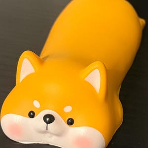 May include: A yellow Shiba Inu dog figurine in a lying down position. The figurine has black eyes, a black nose, and pink cheeks. The ears are white-tipped. The dog is a solid color with a smooth texture.