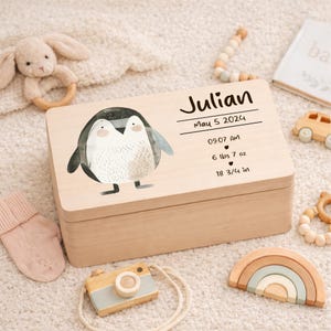 May include: A light wooden keepsake box with a penguin illustration and the name "Julian". The box includes the birth date, time, weight (6 lbs 7 oz), and length (18 3/4 in). Other baby toys are scattered around the box.