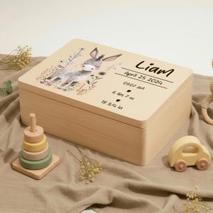 Può includere: Un cofanetto ricordo in legno chiaro con un'illustrazione dipinta di un asino e accenti floreali. Il coperchio è iscritto con il nome "Liam", la data, l'ora, il peso (2,9 kg) e la lunghezza (47,6 cm). Un giocattolo impilabile in legno e un'auto giocattolo sono nelle vicinanze.