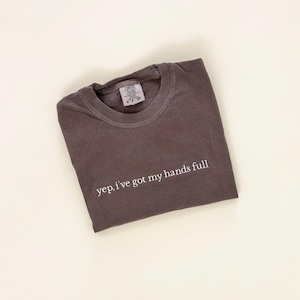 May include: A folded, dark gray t-shirt with the text "yep, i've got my hands full" in white. The shirt has a crew neck and a small tag at the neckline. The shirt is made of a soft material.