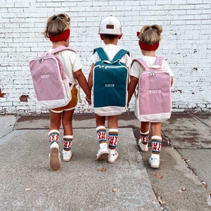 May include: Three children wearing backpacks, socks, and shoes, standing in front of a brick wall. Two children have pink backpacks with white trim, and one has a teal backpack. Each backpack has a name on it.
