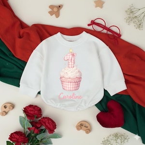 May include: White baby bodysuit with a pink cupcake graphic featuring a number one candle and the name "Caroline". The design includes a pink gingham pattern. The bodysuit is surrounded by decorative elements.
