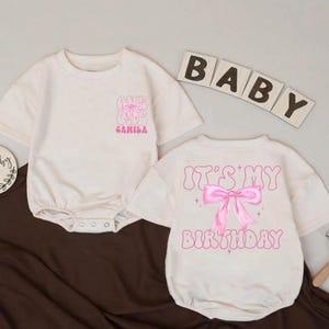 Op de afbeelding: Twee crèmekleurige babyrompers. Eén heeft de tekst "ONE" en "CAMILA" in roze. De andere zegt "IT'S MY BIRTHDAY" met een roze strik-afbeelding. Houten blokletters spellen "BABY".