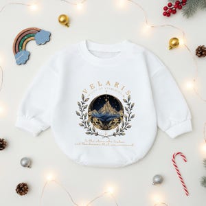 May include: White baby onesie with long sleeves. The front features a circular design with the text "VELARIS CITY OF STARLIGHT" and a mountain scene in blue and gold. Below the design is the text "To the stars who listen and the dreams that are answered."