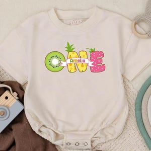 May include: Cream-colored baby bodysuit with the word "ONE" in colorful fruit-themed letters. The "O" is a kiwi, the "N" is a pineapple, and the "E" is a strawberry. The name "Amelia" is on a banner.