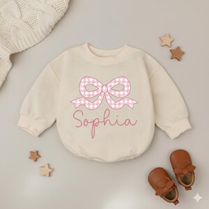 Puede incluir: Body de bebé color crema con un lazo de cuadros rosas y el nombre "Sophia" en cursiva. El body de manga larga se combina con zapatos de bebé de cuero marrón y juguetes de madera con forma de estrella.