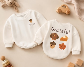 Puede incluir: Dos sudaderas de manga larga de color crema. Una presenta un pequeño gráfico de bellota con el nombre Amelia impreso debajo. La otra muestra la palabra Grateful sobre ilustraciones de un pavo, un trozo de tarta, una bellota y una hoja.