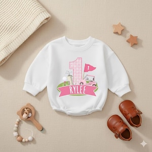 May include: White baby bodysuit with a golf-themed graphic. The design includes a large number 1 in pink gingham, a golf cart, a golf ball, and the name "RYLEE" on a pink banner. Brown leather baby shoes and wooden toys are also in the image.