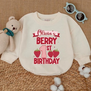 May include: Cream-colored baby onesie with the text "Olivia's Berry 1st Birthday" in red. The design includes strawberries and a checkered number one. A teddy bear and blue sunglasses are next to the onesie.