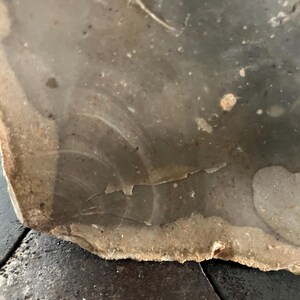 May include: Close-up of a piece of flint. The stone has a light grey and brown colour with a rough, textured surface. The edges are jagged and irregular, with some areas showing signs of flaking.