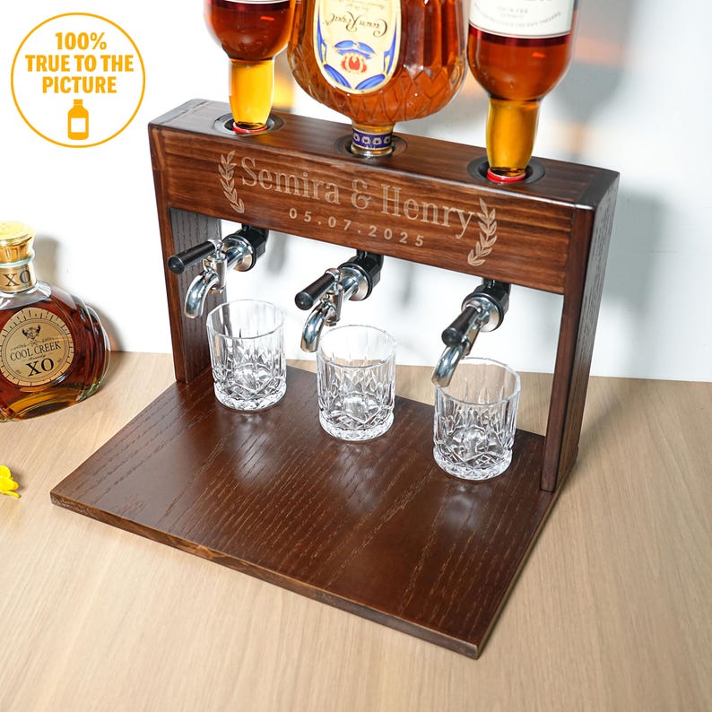 May include: A dark brown wooden liquor dispenser with three spouts, three glasses, and three bottles. The dispenser is engraved with "Semira & Henry" and "05.07.2025". The bottles are filled with amber liquid. The image also includes a bottle of Cool Creek XO.