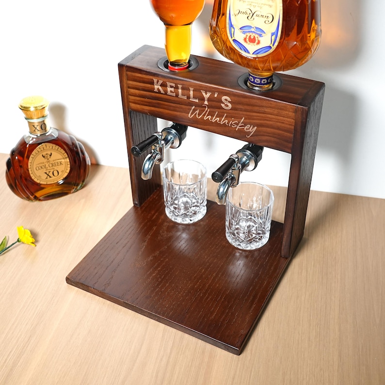 May include: A dark wood whiskey dispenser with two spouts and two crystal glasses. The dispenser has the text "KELLY'S Whhhiskey" engraved on the front. Two bottles of liquor are placed on top of the dispenser.