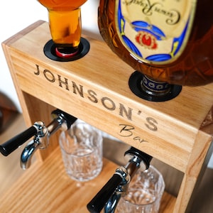 May include: Wooden bar station with the text "JOHNSON'S Bar" engraved on the front. The station has two openings for bottles and two spouts with black handles. Two clear glasses are positioned below the spouts.