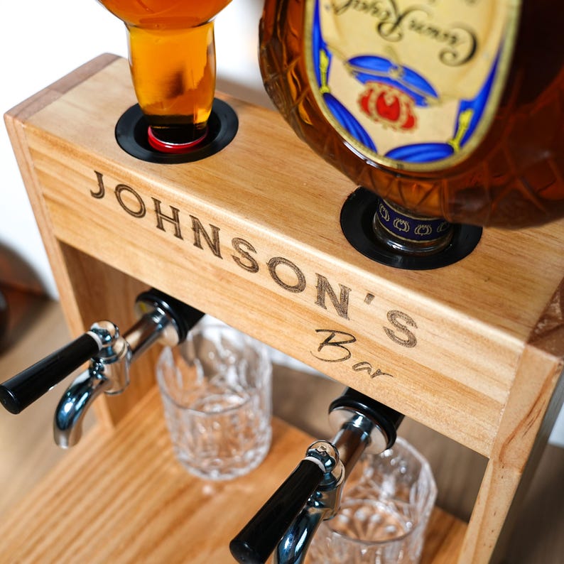 May include: Wooden bar station with the text "JOHNSON'S Bar" engraved on the front. The station has two openings for bottles and two spouts with black handles. Two clear glasses are positioned below the spouts.
